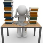 person sitting at a desk with two piles of books with their hands on the head.